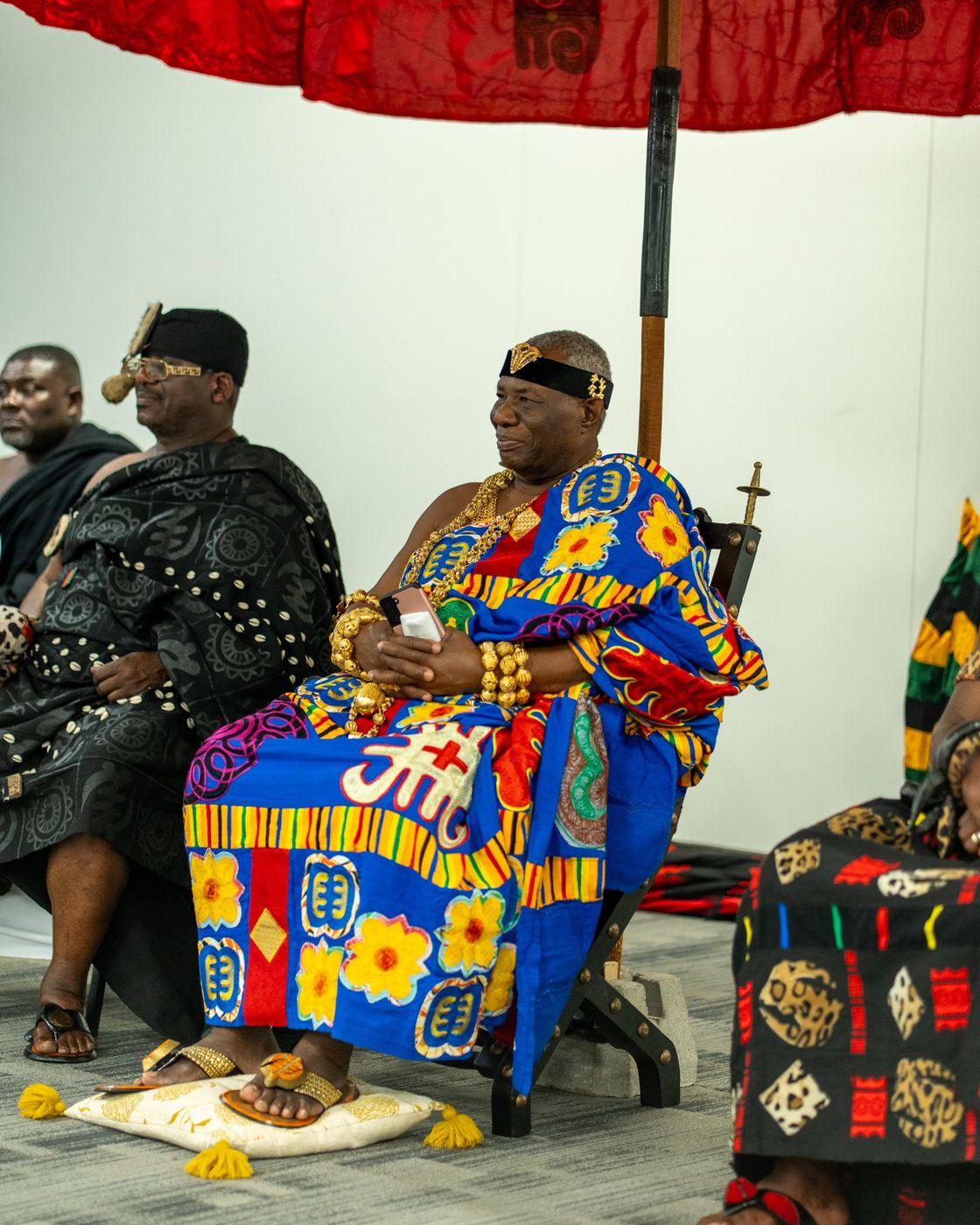 A chief in kente and gold regalia seated beneath a red state umbrella